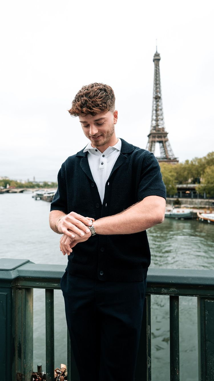 A man, likely the creator, stands casually on a bridge, looking at his watch. The Eiffel Tower is prominent in the background on an overcast day. He is wearing a dark navy cardigan over a white polo shirt and dark trousers. The bridge has a green railing.