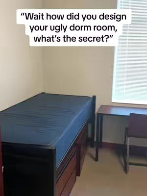 The image shows a plain, functional dorm room. There's a bed with a blue sheet, a desk, and a window with blinds. The walls are empty and cream-colored, and the overall feel is basic and practical.