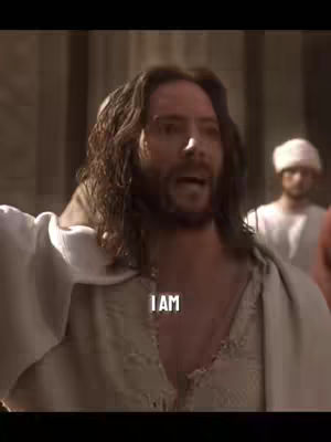 The image shows a medium shot of a man with long brown hair, a beard, and wearing a cream-colored robe. He has an open mouth and appears to be speaking passionately. Overlayed on the image is the text "I AM" in a white, bold font. The background is blurry, with hints of architectural structures and other figures. The lighting is warm and suggests an outdoor setting.
