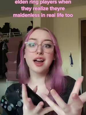 A woman with pink hair and glasses is holding a black controller, with a surprised expression, in front of a cluttered background.