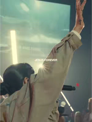 A person with their arm raised, wearing a tan jacket, with a stage-like background with lighting and a person holding a microphone, the text "JESUS FOREVER" is written across the image.