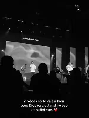 A black and white image taken inside a church, showing a stage with a large screen displaying the text "ALL HAIL KING JESUS". In the foreground, silhouetted figures partially obscure the view of the stage, where several people are standing and some are holding microphones. Overlayed at the bottom is text that reads: "A veces no te va a ir bien pero Dios va a estar ahi y eso es suficiente." and a red heart symbol.