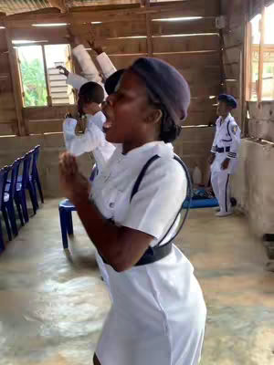A woman in a white uniform and a blue cap is singing in a church during a worship service. Other people are visible in the background with arms raised up.
