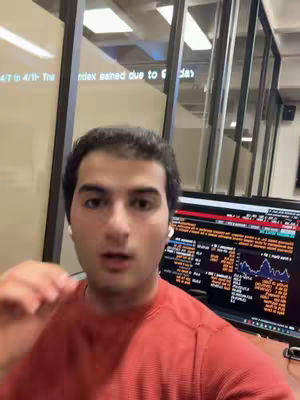A man in a red shirt is centered in the frame against a background of windows and a computer monitor displaying data. The man is looking directly at the camera. The monitor is displaying data and graphs. Overall lighting is bright and natural.