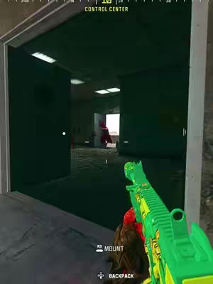 The image shows a first-person perspective from a video game. A green gun is held in the foreground, partially obscuring the lower right. It is pointed down a dark hallway or doorway, revealing a figure in red in the distance. The setting has the feel of warehouse with a control center marked above the doorway.