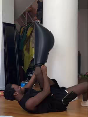 A man lies on the floor with his legs raised to his head, holding one leg with his hands. A large, black, boot-shaped inflated object is held by his other leg.