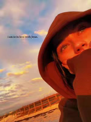 A close-up shot features a person in a brown hooded jacket, partially obscuring their face with their hand. The background shows a cloudy sky with warm sunset colors, and a beach scene with a wooden fence at the bottom edge. Text overlay reads, 'I am so in love with Jesus.'