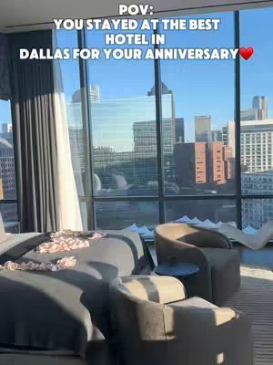 A room with a large glass window showing a city skyline. The room is furnished with a bed covered in a gray blanket and decorated with rose petals, and two beige armchairs. The text overlay on the image reads 'POV: YOU STAYED AT THE BEST HOTEL IN DALLAS FOR YOUR ANNIVERSARY ❤️'.