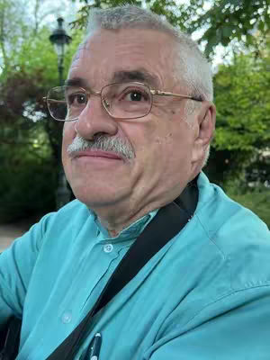 A close-up shot of an older man with glasses and a mustache in a park setting. The man is wearing a teal button-up shirt, and a black strap is visible on his left shoulder. Behind him, blurred trees and a lamppost provide a soft, green background.
