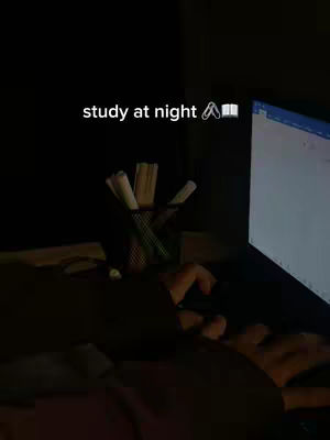 The image shows a dimly lit scene of someone working on a computer at night. The person's arms and hands are visible, typing or interacting with the computer, which has the screen partially illuminated. The background is very dark with a pencil holder and some pencils and pens visible. Text overlayed at the top reads study at night