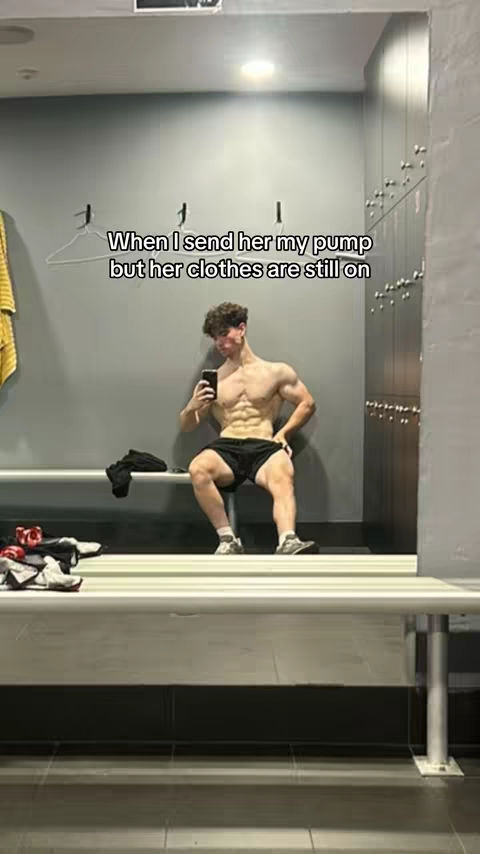 A man with a muscular, bare torso sits on a bench in a locker room, holding a phone and taking a selfie in front of a mirror. Text overlayed on the image.