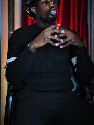 A man in a black outfit is sitting in a chair with his fingers intertwined. He is wearing a cap or durag. The background is a red curtain. The man is looking upwards.