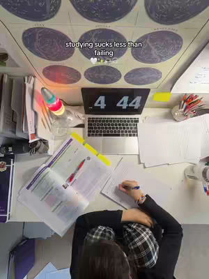 An overhead shot of a study desk with a laptop displaying 4:44, open textbooks, notes, a lava lamp, and supplies. The person is wearing a plaid hat.