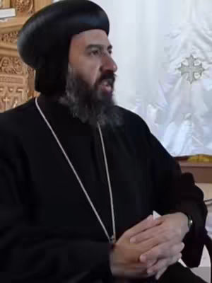 A medium shot features a middle-aged man, likely a religious leader, wearing a black robe and head covering. He has a full beard and is looking slightly off-camera to the left. The background is softly lit, with a glimpse of architectural details on the left and a white, decorative pattern on the right. The composition has a subdued, respectful tone.