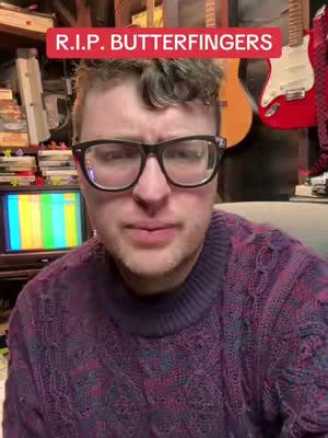 A man with glasses is positioned in the center, looking at the camera. Above him is a red banner with white text, and the background includes guitars and shelves.