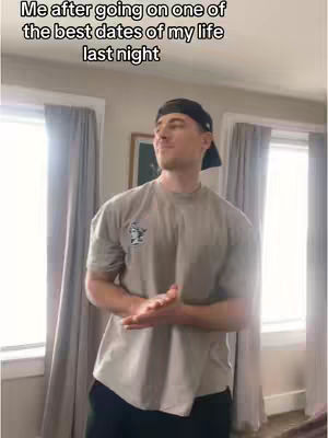 A man in a gray shirt and black cap stands indoors, smiling slightly with his hands clasped. The background includes windows with curtains and a picture frame.