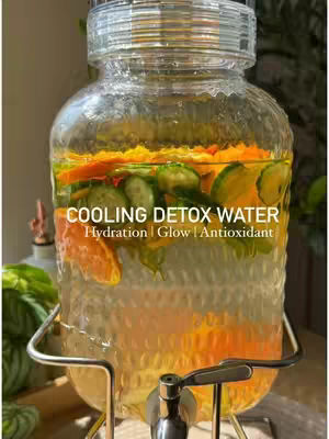A close-up shot of a large glass beverage dispenser filled with clear water, sliced oranges, lemons, and cucumbers. The bottom half of the dispenser is filled with a lighter colored liquid, and a metal spigot is visible at the bottom. The dispenser sits on a metal stand, and leafy green plants and other objects are blurred in the background. Text overlays the image.