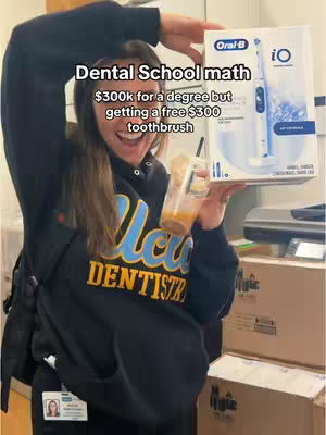 A smiling woman holds a box of an Oral-B iO toothbrush, a cup with a straw, and wears a dental school sweatshirt. Text overlayed includes: 'Dental School math, $300k for a degree but getting a free $300 toothbrush.' Behind her are boxes in a room, she appears to be standing in.