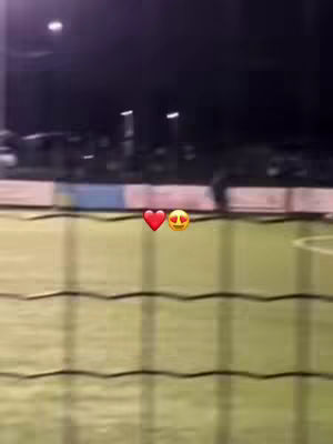 A blurry photo of a soccer field at night. The field is green, and the background is dark. There are lights in the distance. In the foreground, there's a fence. There are emojis of a heart and a face with hearts for eyes in the image.