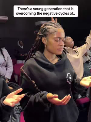 A woman with braided hair is centered in the frame, eyes closed, with hands raised in a worshipful gesture. Other people are visible in the background, also with hands raised in prayer. The setting appears to be a church or similar gathering space. The image has a warm, indoor lighting with overall positive vibe.