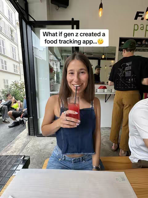 A woman with long brown hair is sitting at a table in a cafe or restaurant holding a pink drink and smiling. She is wearing a blue denim top. A text overlay in a white box reads: "What if gen z created a food tracking app...?" There are other people in the background, including a person with a green cap.