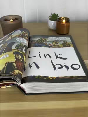 An open comic book with text overlayed, next to a candle and a small plant, on a wooden table. The comic book is open to a page with illustrations. The text reads "Link n bio" in a handwritten style. A candle is lit to the left. A small plant in a white pot and a small lit candle are in the background.