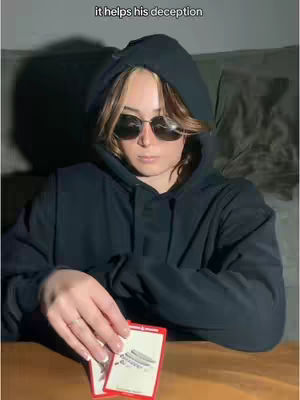 A person with light brown hair wearing sunglasses and a black hoodie is holding two cards in their hands. The text overlay says, “it helps his deception”. The background appears dark with hints of a room setting.