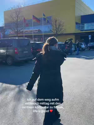 A person is walking away from the camera in an outdoor shot. The person is wearing a long, dark coat. The background includes an IKEA store with flags waving. Text is overlayed on the image.