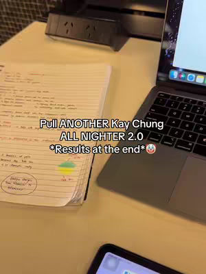 A top-down shot captures a study setup. A laptop is open on the right, displaying a brightly lit screen. To the left, a notebook is open, with handwritten notes and diagrams. Beneath the notebook, the corner of a phone is visible. The desk surface is a light color, and a power outlet is out of focus in the background.