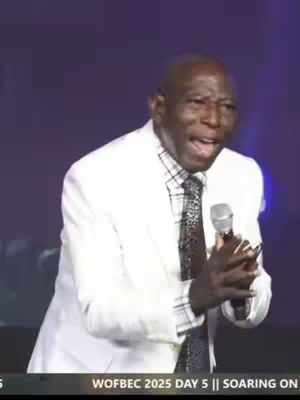 A man in a white suit is holding a microphone and speaking passionately. The background has a blurry purple color.