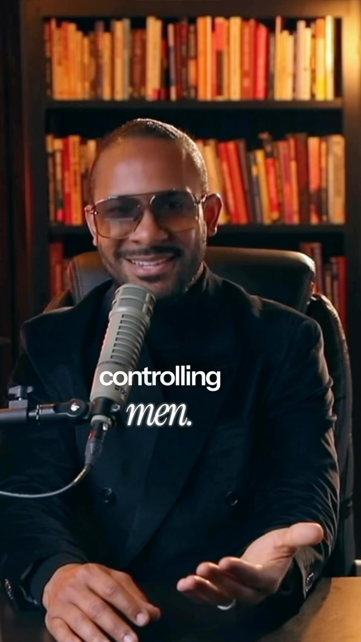 A man with dark skin wearing glasses and a black outfit is speaking into a microphone. The background is an out-of-focus wooden bookshelf filled with books.