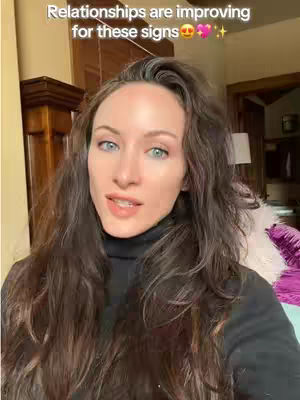 A woman with long, wavy brown hair and fair skin is looking directly at the camera with a slight smile. She is wearing a black turtleneck. Behind her, the background shows a glimpse of a room with a wooden cabinet.