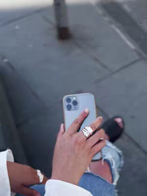 A person is shown holding a smartphone with both hands. Their hands are prominently in the foreground. The phone is angled towards the viewer, with the screen facing away. The person is wearing a ring on their index finger. The background appears to be a paved area, and the person is seated, with their legs and feet visible. The overall lighting suggests an outdoor setting.