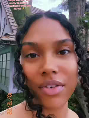 A close-up shot of a woman with dark curly hair, speaking to the camera while set against a blurred background of a house and greenery. Text overlays the image, emphasizing a motivational message.