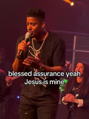A man in a black t-shirt holds a microphone and sings on stage. A woman is visible on the right side of the frame. The text overlay reads "blessed assurance yeah Jesus is mine". The background shows a stage with lighting.