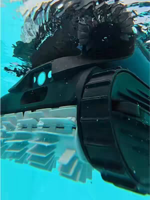 An underwater shot of a robotic pool cleaner. The cleaner is black and has large, rugged treads with white brushes. It is partially submerged in blue water, with ripples visible on the surface.