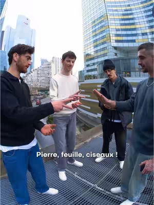 The image features four men on a rooftop, playing 'rock, paper, scissors'. They are dressed in casual attire, with one person clearly making the 'scissors' gesture, while the others are in different poses. The background includes a cityscape and a tall modern building with reflective glass.