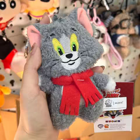 A hand holding a Tom and Jerry plush keychain. The Tom cat character is gray with white and pink accents, wearing a red scarf. The background is filled with other plushies and merchandise, with a partial view of a product tag.
