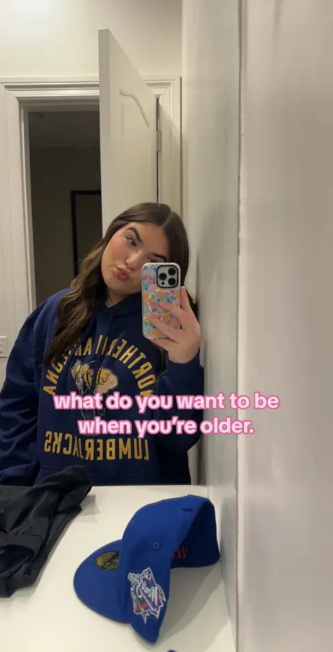 A selfie taken in a bathroom mirror with a woman holding a phone. The woman is wearing a dark blue hoodie with yellow text. In the foreground, a dark blue cap rests on a white surface.