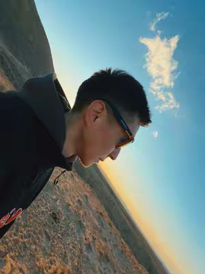 A young man wearing sunglasses and a hoodie is seen in profile against a backdrop of a blue sky and rolling hills. The composition is angled, creating a dynamic feel. The lighting suggests either sunrise or sunset, with warm tones across the landscape and cool blues dominating the sky.
