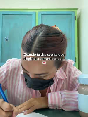 A woman with dark hair and a black face mask is writing on a paper. She is wearing a pink and white striped shirt with the sleeves rolled up. In the background, there are two green cabinets.