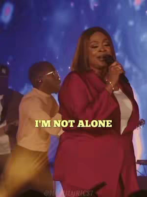 A woman with brown hair, wearing a red dress and holding a microphone, appears to be singing on stage. Beside her, a man wearing glasses and a light-colored shirt stands out of focus. The overlay text on the image is 'I'M NOT ALONE' in a gold color.