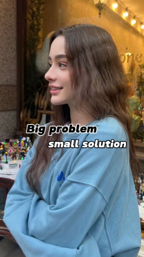 The image features a young woman in a light blue sweatshirt with the text "Big problem small solution" overlaid on top. She's seen from the side, looking towards the left, against a somewhat blurred background suggestive of an outdoor setting with what looks like a store or storefront. The lighting appears natural, and the overall aesthetic is clean and modern.