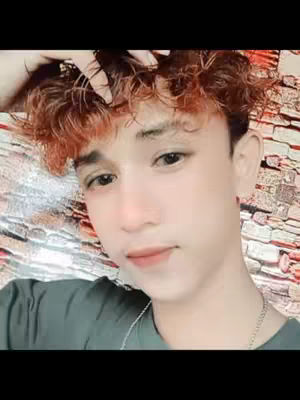 A close-up shot of a young man with light skin and curly reddish-brown hair. He's looking slightly off-camera with a subtle smile. He wears a dark green shirt and a silver chain. The background appears to be a textured wall with shades of white, gray and brown.