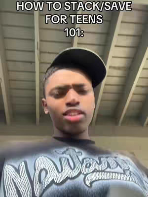 A young man with a black cap and a black shirt with white text looks directly towards the camera, with what looks like a silver ceiling in the background.