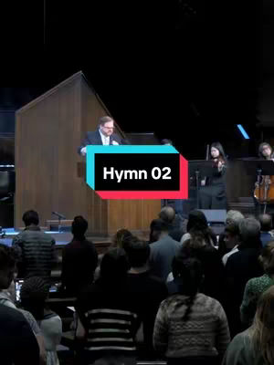 A vertical image taken in a church. An audience of people is facing the front. A man is standing at a podium at the front, and a lady is playing the violin. A graphic with the words "Hymn 02", blue and red, is placed in front of the man at the podium.
