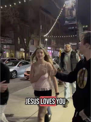The image captures a street scene at dusk. A young woman, center and facing slightly toward the viewer, walks along a sidewalk with a slight smile, wearing a pink top and black shorts. To her right is a man in a hoodie. The text overlay 'JESUS LOVES YOU' appears in a red-bordered box. 