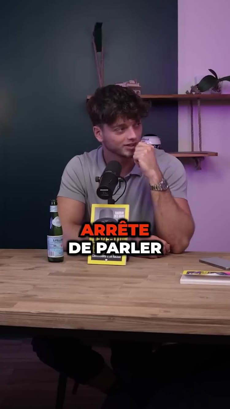 A man sits at a wooden table with a microphone. He wears a grey polo shirt. In front of him is a book with a yellow cover and the words 'ARRÊTE DE PARLER' in large, bold, white letters with a red outline.