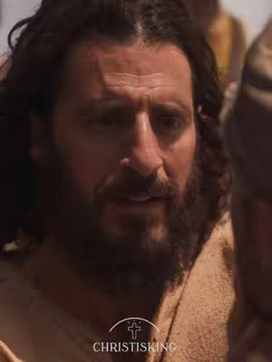 The image is a close-up of a man with dark, wavy hair and a full beard looking towards the right. His skin has a warm tone, and he seems to be in a scene with other people, some of whom are partially visible. The setting appears to be outdoors or in natural lighting. In the lower center of the frame, a logo with text is visible, further contextualizing the image.