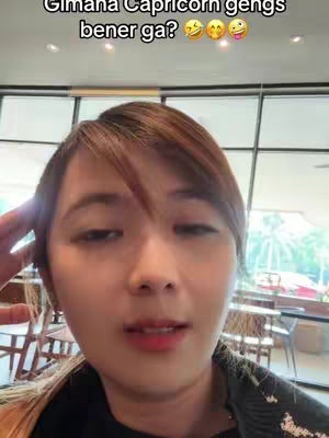 A selfie-style video with a woman's face as the central focus. She has short brown hair and is looking towards the camera with a somewhat serious expression. The background consists of a room interior with window views and tables and chairs, suggesting a public space. A text overlay at the top says, "Gimana Capricorn gengs bener ga?" and includes emoji of smiling faces with different expressions.